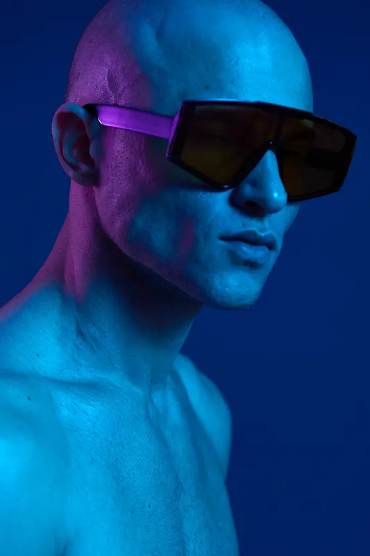 Stylish man with sunglasses posing against a vibrant blue and purple background, showcasing modern fashion and artistic expression in color light.