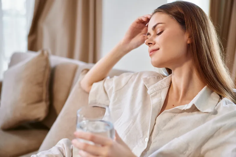 Woman on sofa holding a glass of water, relaxing at home with eyes closed in a calm portrait. Soft natural light, casual shirt, cozy living room and peaceful moment.