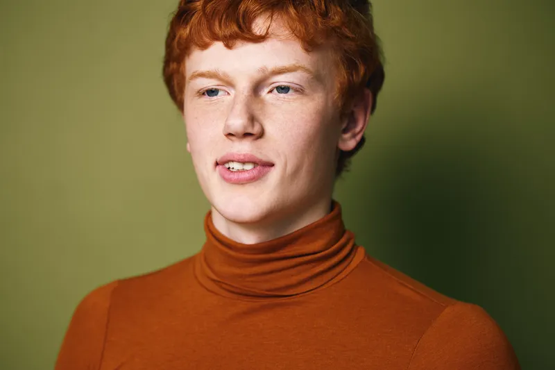Young man with red hair and fair skin wearing a brown turtleneck sweater against plain green background. Casual portrait of thoughtful male with freckled face and relaxed expression.