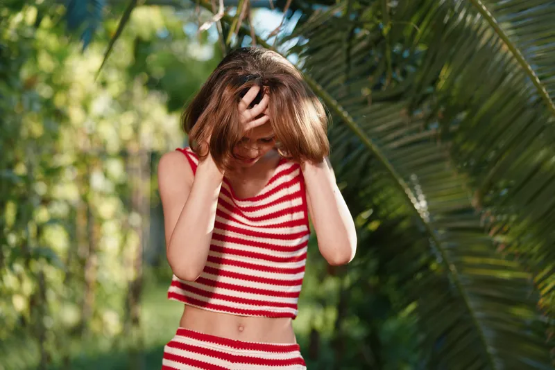 A young woman wearing a red and white striped crop top stands outdoors in a sunlit garden, her hands brushing hair from her face, creating a casual, carefree mood.
