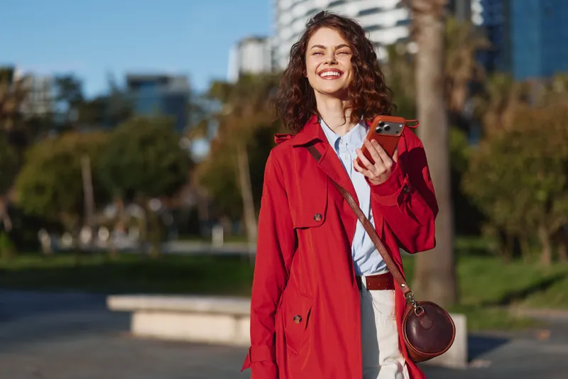 A smiling young woman in a red coat holds a smartphone outdoors in a sunny urban park with buildings in the background. Concept for technology and lifestyle.