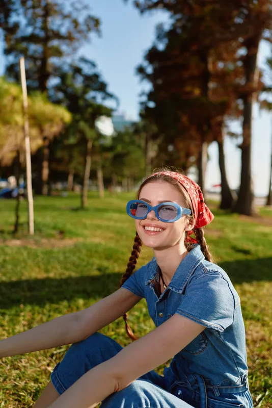 Lifestyle woman in modern boho-western denim street style wearing blue sunglasses and red bandana, smiling outdoors in warm film color light with natural green background.