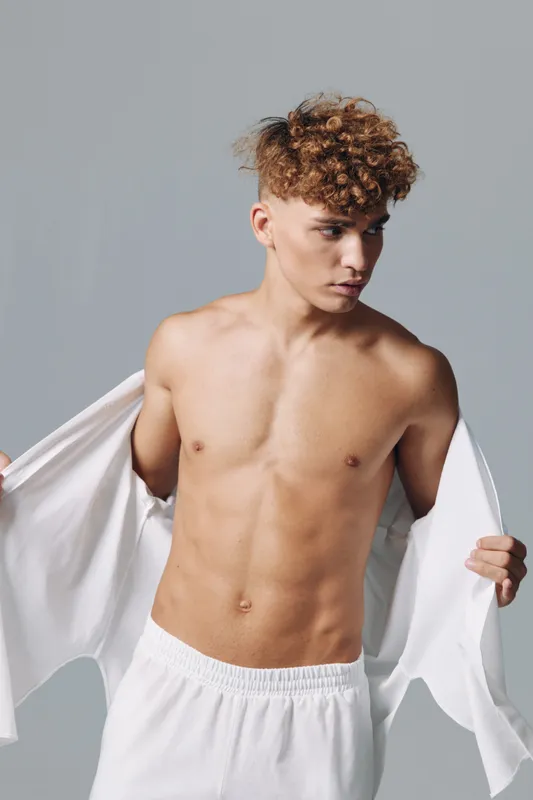 Young man with curly hair and bare chest wearing white pants and holding open white shirt on gray background. Fitness, casual style and confident pose with focused expression.