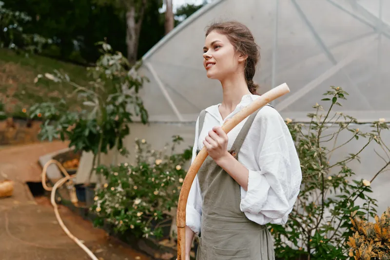 Young woman in light clothing smiling and holding a gardening tool in a green environment, symbolizing connection with nature and sustainable living