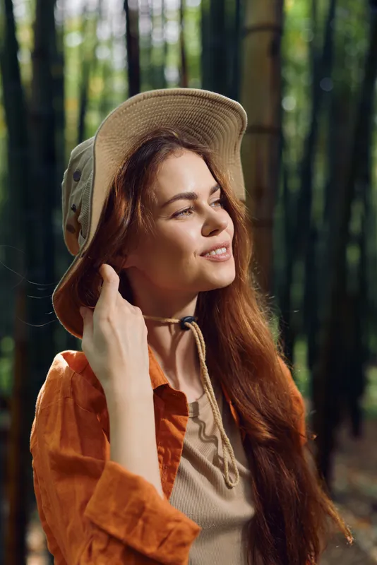 Woman in hat standing in bamboo forest, portrait smiling and enjoying sunlight. Redhair model outdoors wearing casual shirt, nature scene, relaxed expression and warm mood.