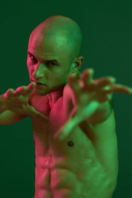 Shirtless man posing dramatically against a dark green background, showcasing strength and intensity. The image captures a captivating, bold expression in color light.