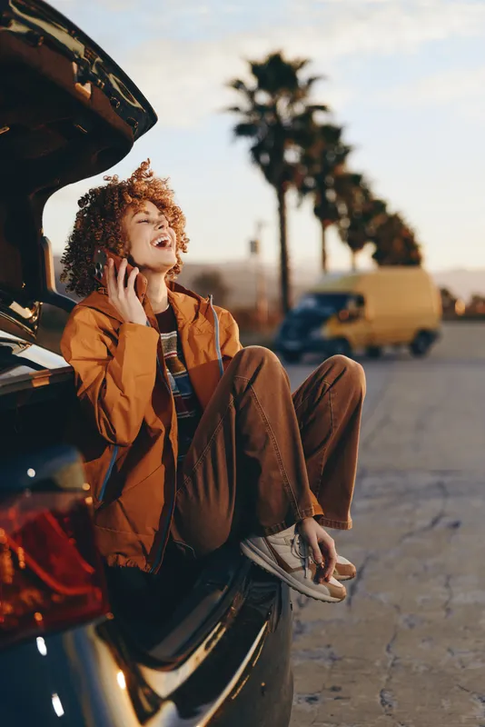 Woman lifestyle by open car door, curly haired woman sits outside, smiling and holding smartphone to ear, warm sunset light, street parking, casual jacket jeans ready for trip outdoors