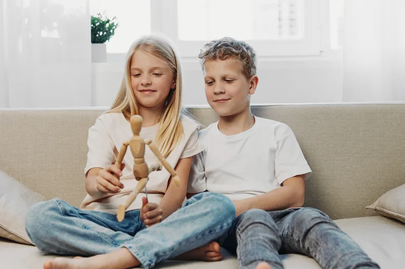 Two children sitting on couch playing with wooden mannequin, smiling and enjoying creative activity in bright living room with natural light and casual clothes.