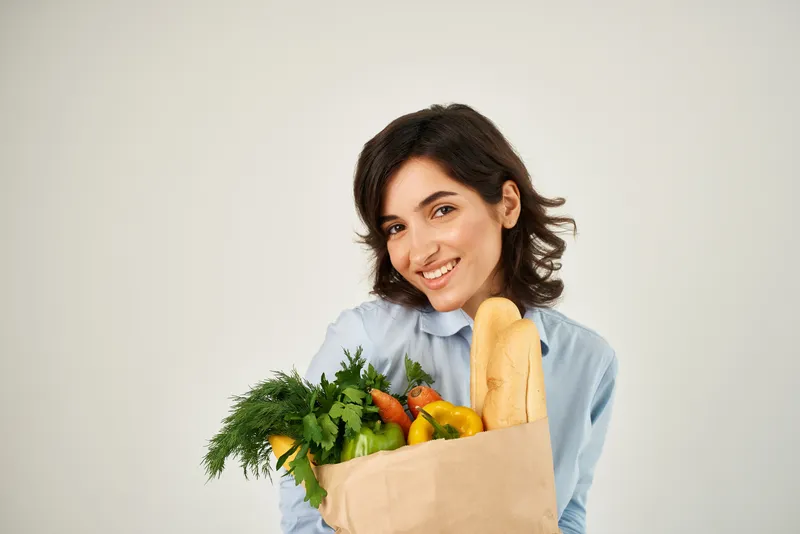 Симпатичная женщина с сумкой продуктов и овощей на светлом фоне, олицетворяющая здоровое питание