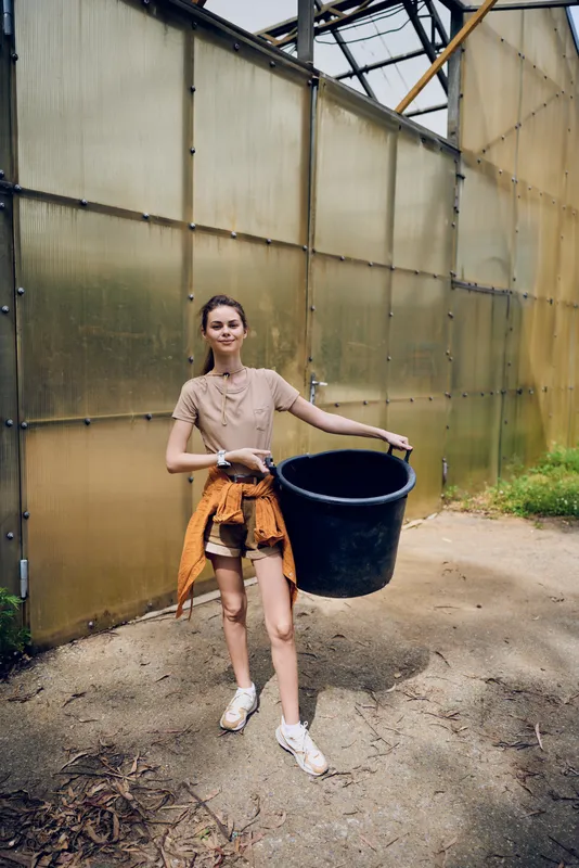 Woman greenhouse tub gardening container casual standing sneakers, holding a large black tub for planting and soil, young female gardener in jacket tied on gravel path by translucent panels.
