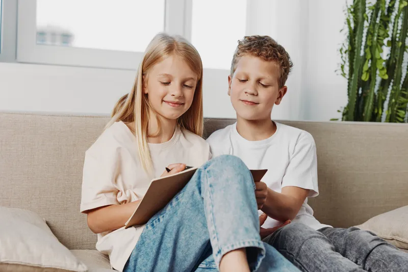 Two smiling children sitting on sofa at home, girl writing in notebook while boy watches. Casual clothes, natural light, calm family moment with focus on learning and creativity indoors.