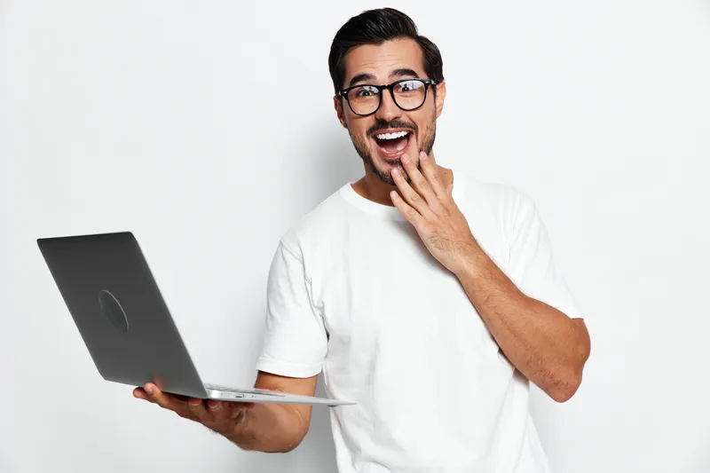 Smiling man with glasses holds laptop while looking at the camera, expressing surprise and joy, isolated on plain solid white background. Technology lifestyle concept.