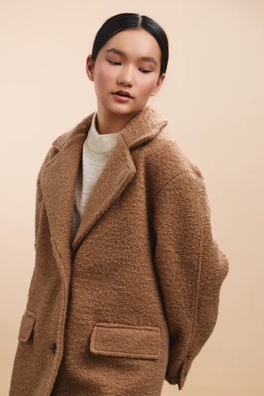 asian woman wearing a tan textured coat stands in a soft studio backdrop, exuding calm confidence and elegant style, a minimalist fashion moment captured with warmth.