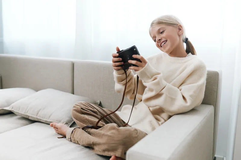 Authentic lifestyle scene of a student at home, relaxed after school, playing with a handheld device, cozy sweater, casual pose, everyday joy and focus on leisure.