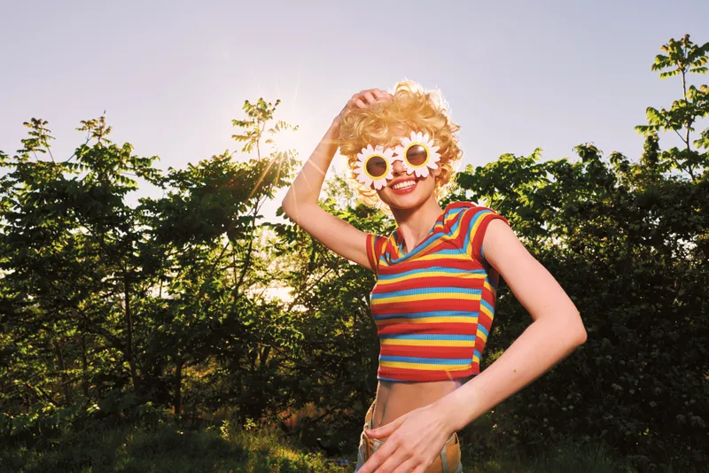 A playful woman in a rainbow striped top and flower sunglasses enjoys a sunny outdoor scene, capturing retro film vibes and a carefree summer mood. with a dreamy cinematic feel.