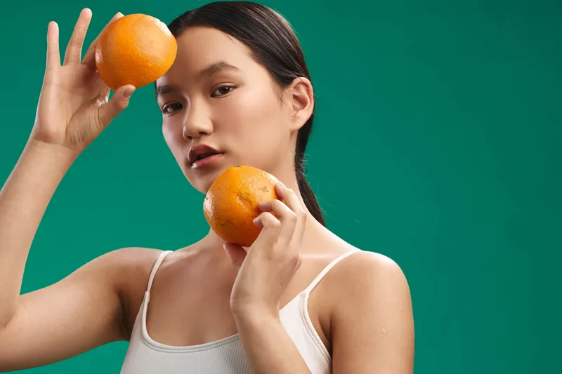 asian woman holds vibrant oranges in a studio, facing camera with a soft smile. bright, clean setting emphasizes fresh, healthy lifestyle and contemporary aesthetic.