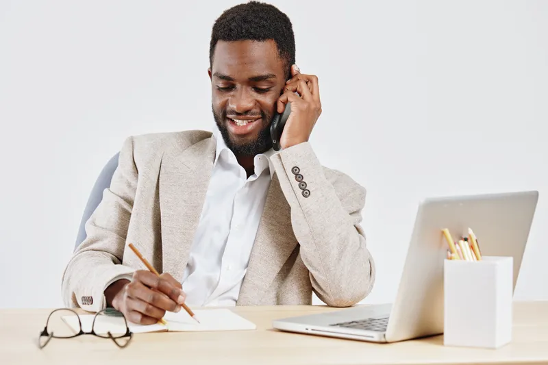 Confident young man in beige blazer smiling while talking on mobile phone at workspace, using laptop and taking notes, in modern office with bright lighting and minimal decor, business communication c