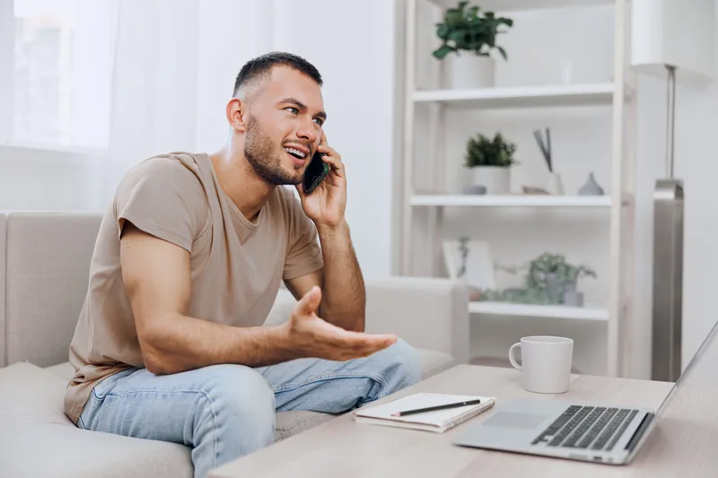 Engaging man on phone, smiling while discussing ideas at home office. Stylish living room enhances the contemporary work-from-home vibe.