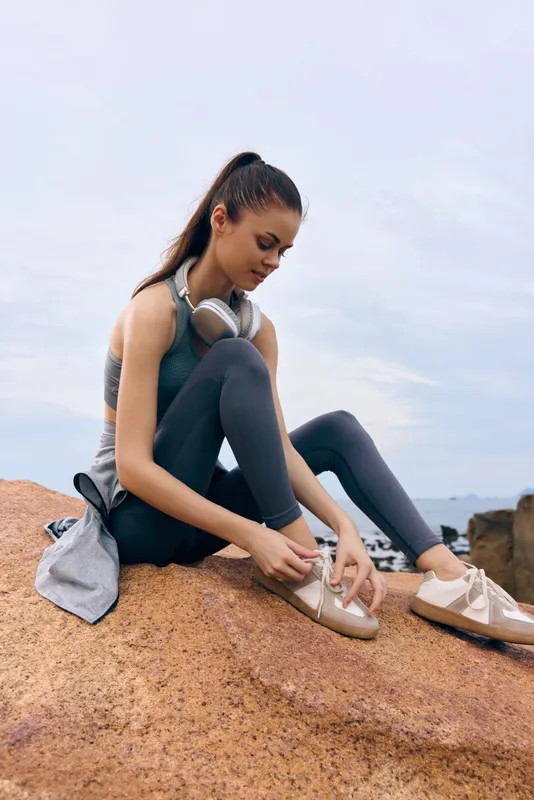 Young female athlete tying shoelaces on rocks, showcasing an active lifestyle in sportswear, emphasizing fitness, determination, and outdoor adventure