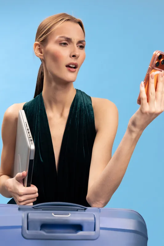 Young woman holding laptop and smartphone stands behind blue suitcase on light blue background. She looks surprised and focused, concept for travel, technology, communication, and business trip.