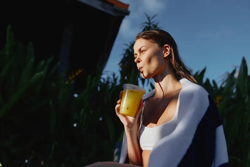 Young woman with wet hair wearing white top and towel drinking juice from plastic cup with straw outdoors. Concept of refreshment, lifestyle, health, and relaxation in natural sunlight.