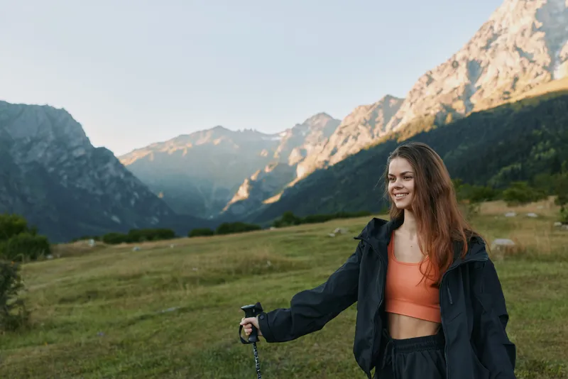 A cheerful woman hiker enjoys an outdoor moment in a mountain meadow. She smiles warmly, wearing a jacket and orange top, holding a trekking pole as sunlight highlights the alpine scenery.