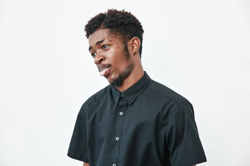 Young black man with dark skin wearing black shirt making funny face with tongue out on white background. Casual expression, studio portrait of African ethnicity male with short hair.
