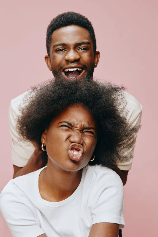 Happy young couple expressing joy with playful facial expressions against a pastel pink background, showcasing vibrant emotions and casual clothing