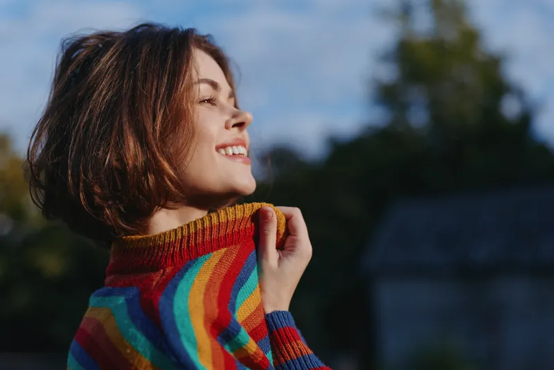 Smiling woman portrait outdoors in profile wearing a rainbow sweater, short hair and closed eyes enjoying warm sunlight, colorful knitted pullover and relaxed happy mood in nature.