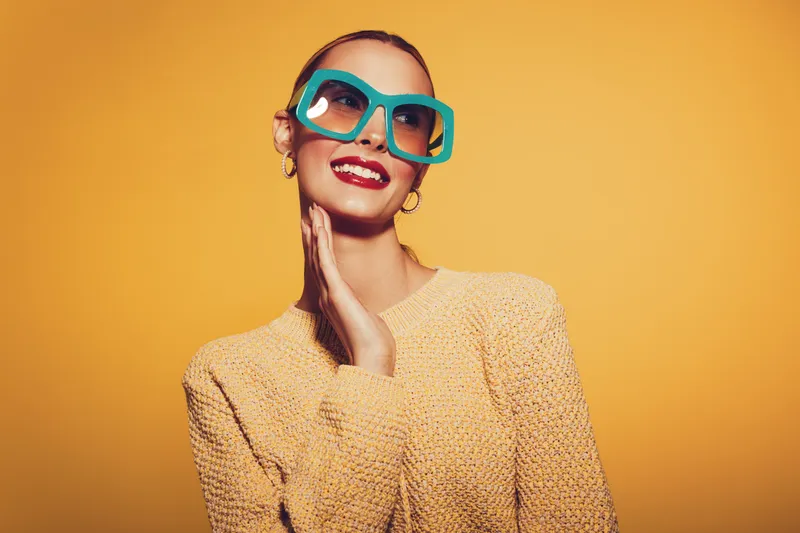 Smiling woman in oversized turquoise sunglasses and yellow sweater poses against bright yellow background. Fashion portrait with vibrant colors and joyful mood, concept of style and confidence.