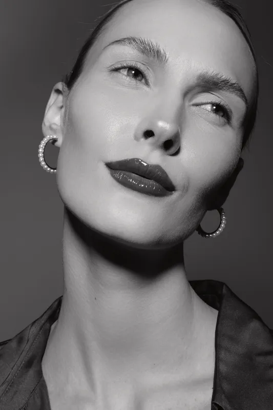 Close-up portrait of a light skinned Caucasian woman with slicked hair and pearl hoop earrings. Studio black and white photo with confident facial expression and elegant style.