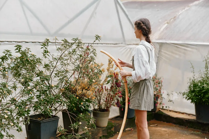 Young woman caring for plants in a greenhouse, wearing a light blouse and shorts, surrounded by colorful flowers and greenery, representing nature and sustainability