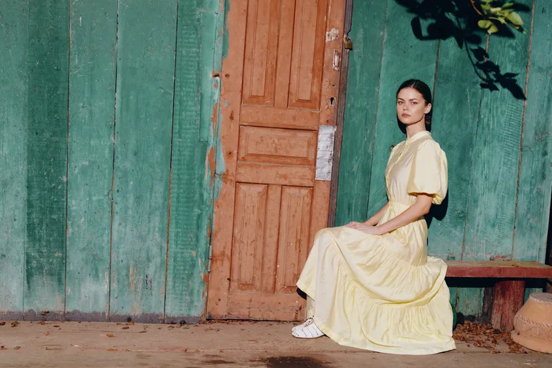 Portrait of a woman in a yellow dress outdoors, sitting by a wooden doorway on a turquoise rustic wall, calm solitary mood, soft daylight, simple vintage setting with elegance and quiet ambiance,