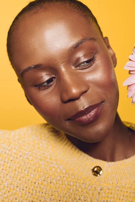 Close-up of happy black woman with dark skin wearing yellow sweater looking down. Bright yellow background and soft natural makeup create warm, positive mood and beauty concept.