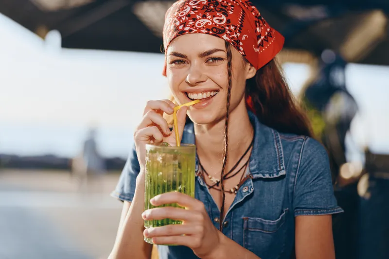 Lifestyle woman with modern boho-western denim street style drinking a green smoothie, smiling with a red bandana and natural film color tones in bright outdoor setting.