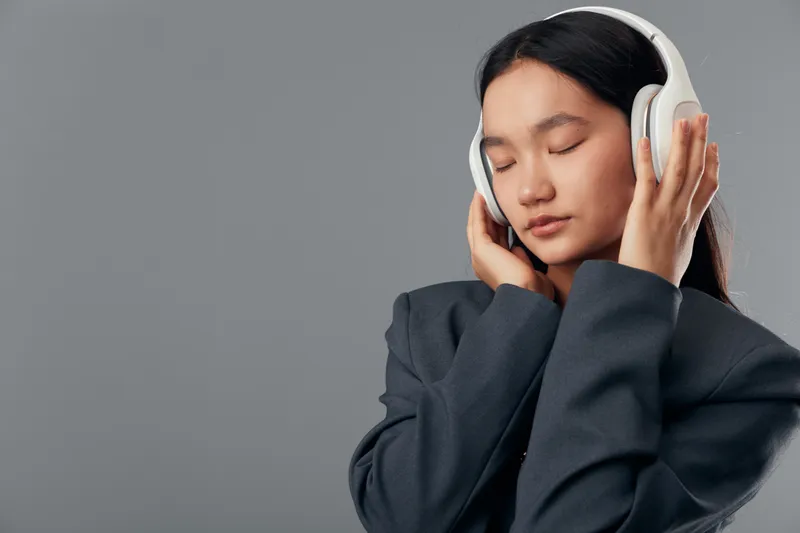 Woman with closed eyes wearing headphones and dark jacket listening to music relaxed and peaceful on gray background in studio. Young female enjoying sound and calm moment.