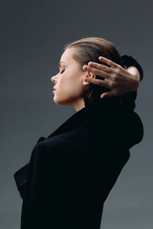 Profile of a thoughtful woman with closed eyes in black clothing against gray background, expressing calm and introspection.