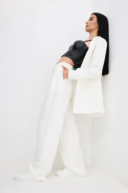 tall woman in white suit leans on wall wearing black top, stylish outfit with long dark hair, minimalistic bright background, modern fashion portrait, trendy look