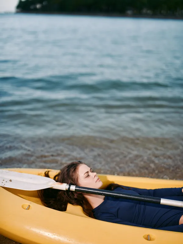 A person lies in a yellow kayak on a calm lake, resting with an oar across the chest, creating a serene, focused moment of outdoor leisure and quiet relaxation.