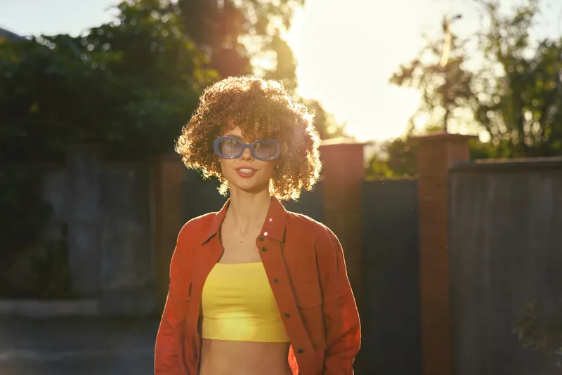 Smiling woman with curly hair wearing a bright yellow crop top and orange jacket outdoors, sunlight bathes the scene, casual lifestyle vibe, warm confident mood