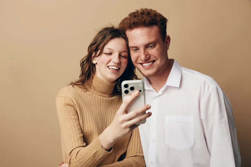 Smiling young couple takes selfie with smartphone on beige background. Casual clothing, happy mood, close relationship, technology use, modern lifestyle, digital communication concept.