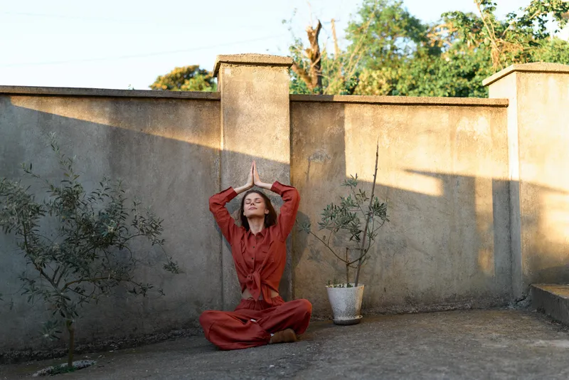 A woman in a red jumpsuit sits cross-legged on the ground against a weathered wall, hands on her head, conveying stress, burnout, and contemplation