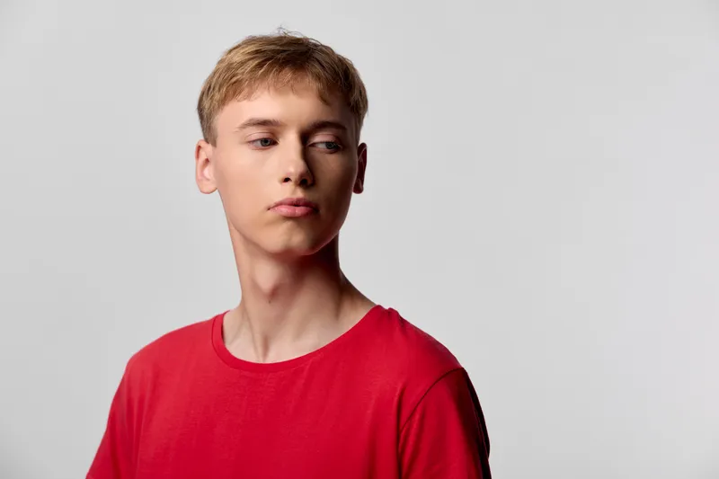 Red shirt, young male in a studio wearing casual fashion, looking away with a neutral expression, minimalist backdrop, clean lighting, contemporary style and subtle mood.