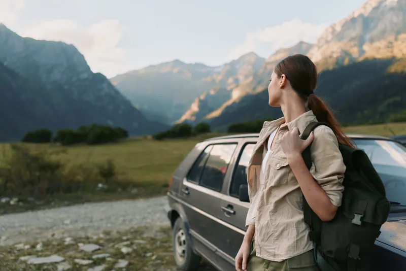 Young woman with long brown hair stands next to a car in a mountain valley looking at the distant peaks while holding a backpack over her shoulder.