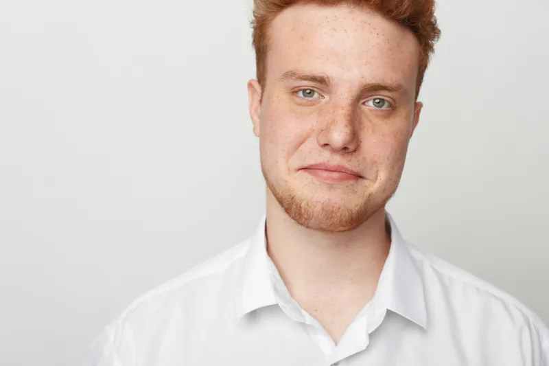 Redhead man with beard and blue eyes wearing white shirt looks at camera with slight smile. Studio portrait on light background. Concept of confidence, casual style, and natural appearance.
