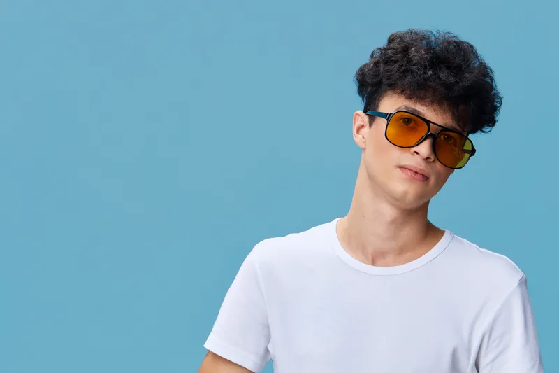 young man wearing yellow sunglasses and white t-shirt posing against light blue background with casual confident expression and curly hair.