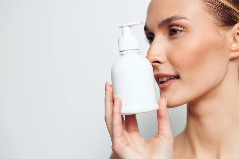 A blonde woman in a minimal studio holds a white pump bottle near her face, conveying a calm skincare moment and clean beauty in a bright, simple setting.