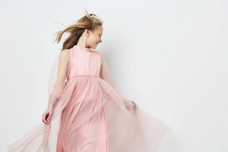 Young girl in a flowing pink dress, smiling joyfully, against a soft pastel background, embodying happiness and freedom in fashion