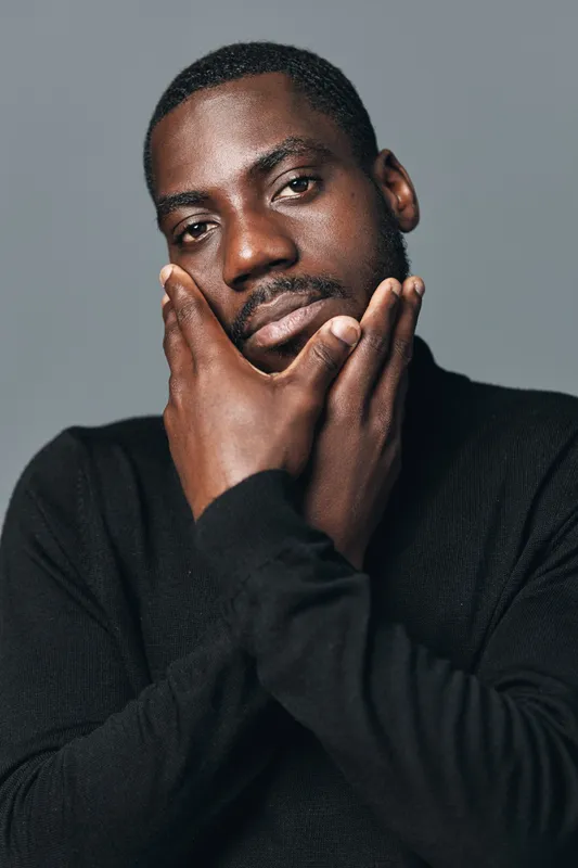African man with dark skin in creative conceptual visual style posing with hands on face wearing black sweater against gray background in studio portrait photography.