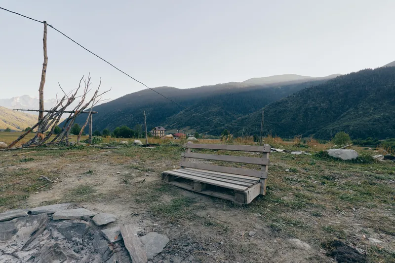 Bench, mountain, field, fence, rural, landscape. Empty wooden bench on a grassy field near rustic fence posts and a stone firepit, overlooking distant mountain slopes and a calm valley panorama.